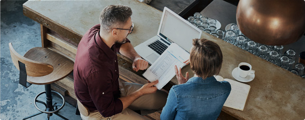 Two professionals in a casual meeting, reviewing documents and using a laptop.