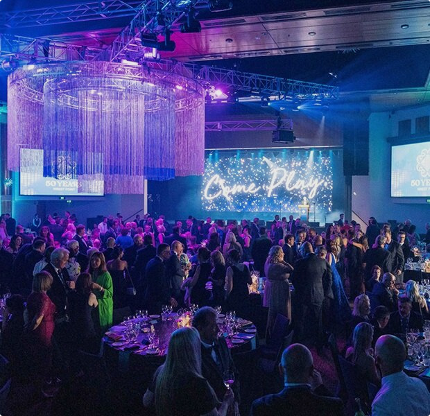 Elegant gala event scene with guests in formal attire, dining under blue lights and large chandeliers.